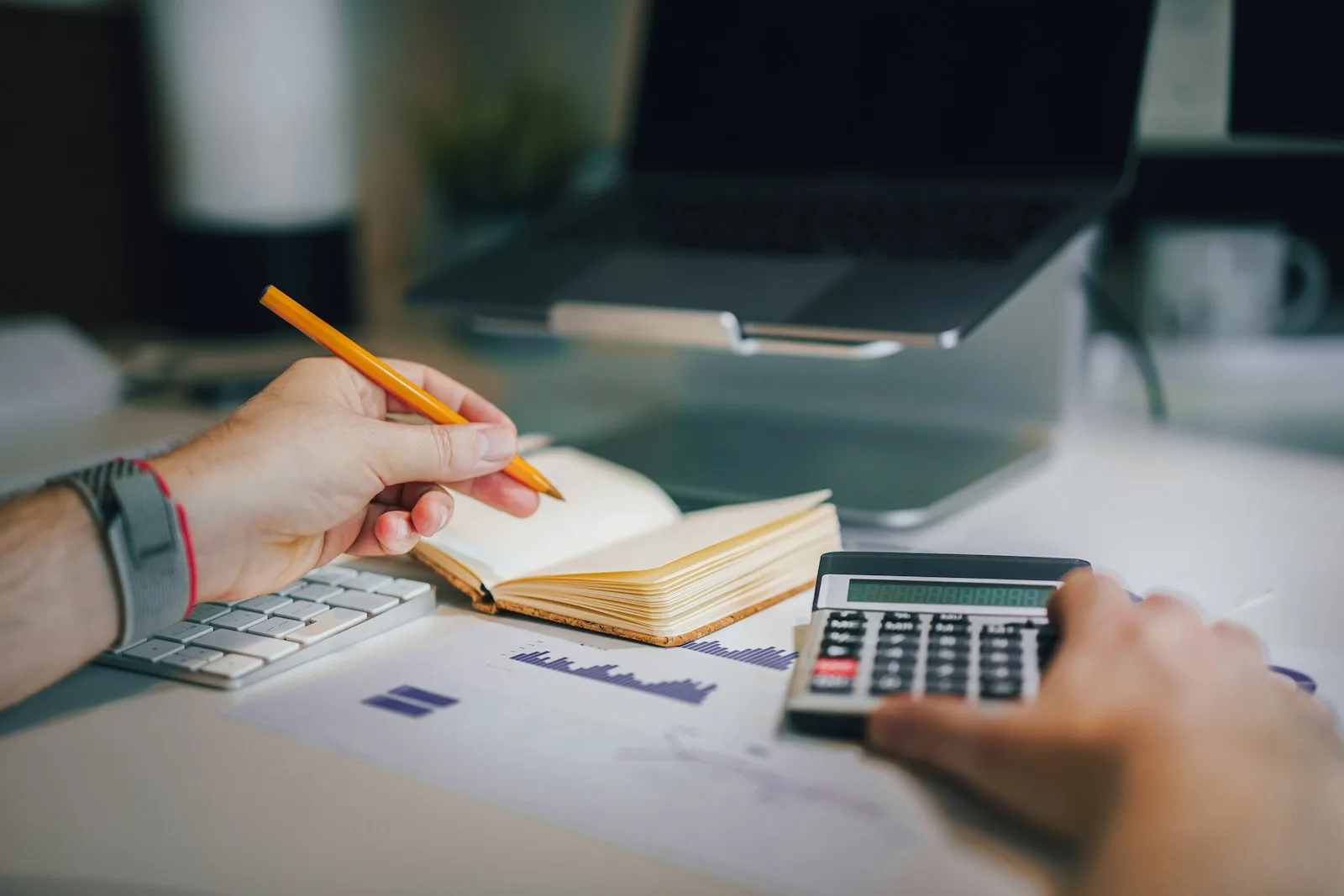 Hands using a calculator next to a notebook, charts, and a laptop on a desk, representing working out the real cost of an AI receptionist