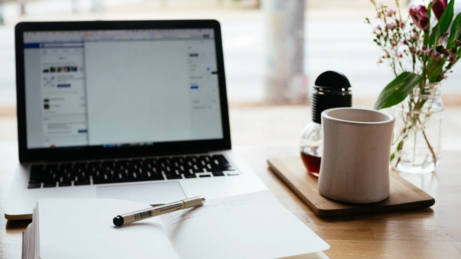 A laptop and an open notebook with a pen on a wooden desk, representing the work of writing and refining an AI receptionist prompt