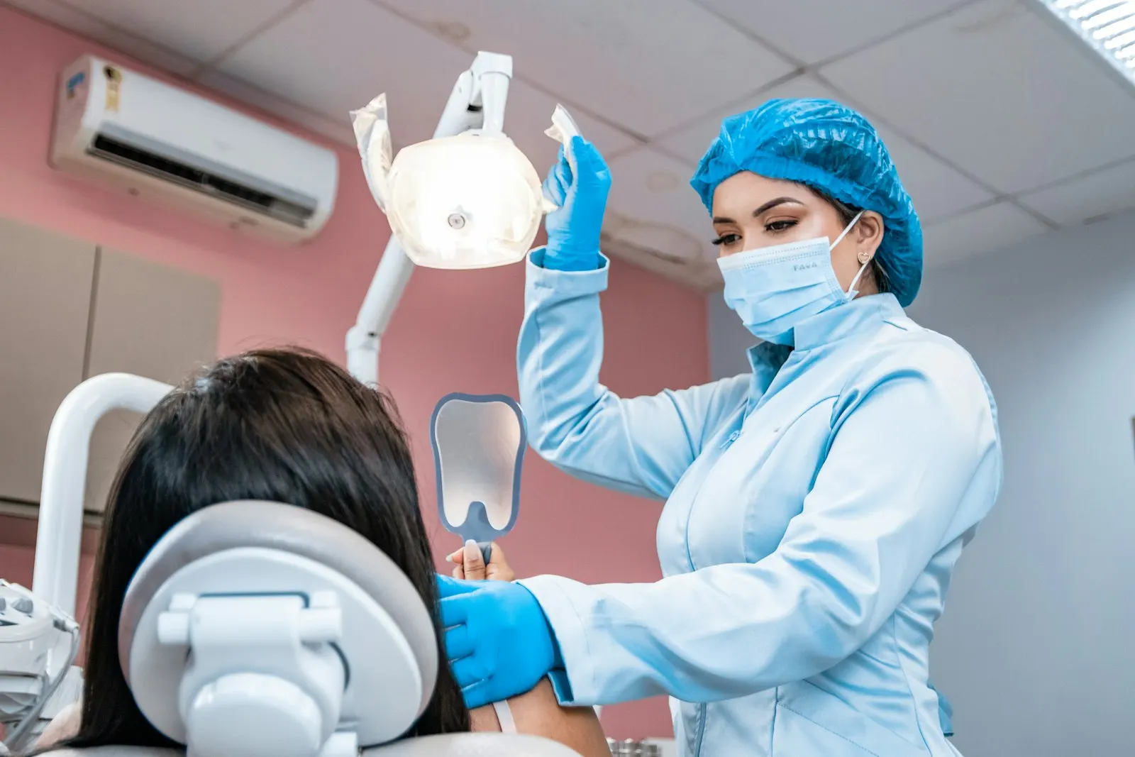 A masked dental professional in scrubs adjusting the overhead light and holding a dental mirror beside a patient reclined in the treatment chair