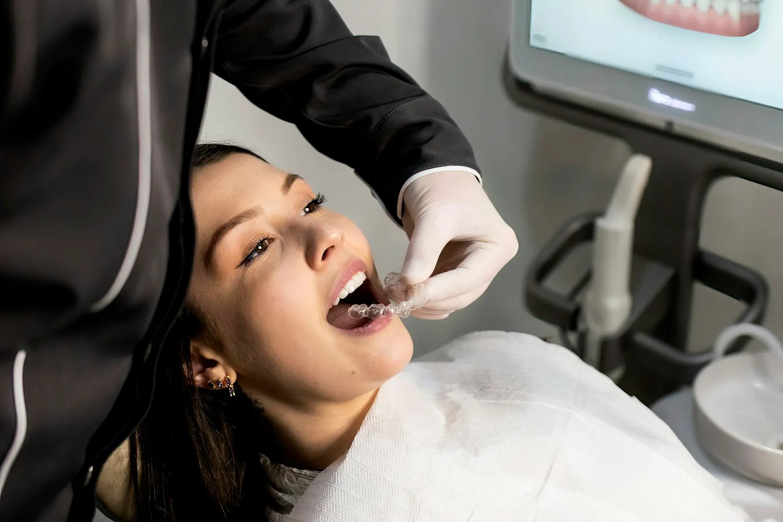A dentist fitting a clear dental aligner for a patient reclined in the treatment chair, with an intraoral scan of the teeth on the screen behind them