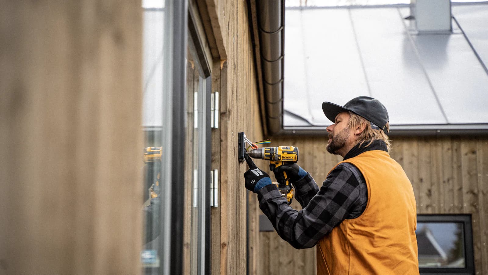 An electrician in a black cap and orange work vest installing an exterior outlet on a wooden house with a cordless drill, both hands on the job while the phone rings elsewhere