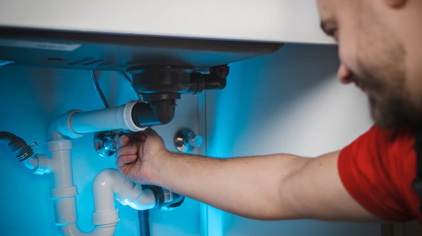 A plumber reaching under a sink to repair the drain trap and supply lines, both hands occupied inside the cabinet